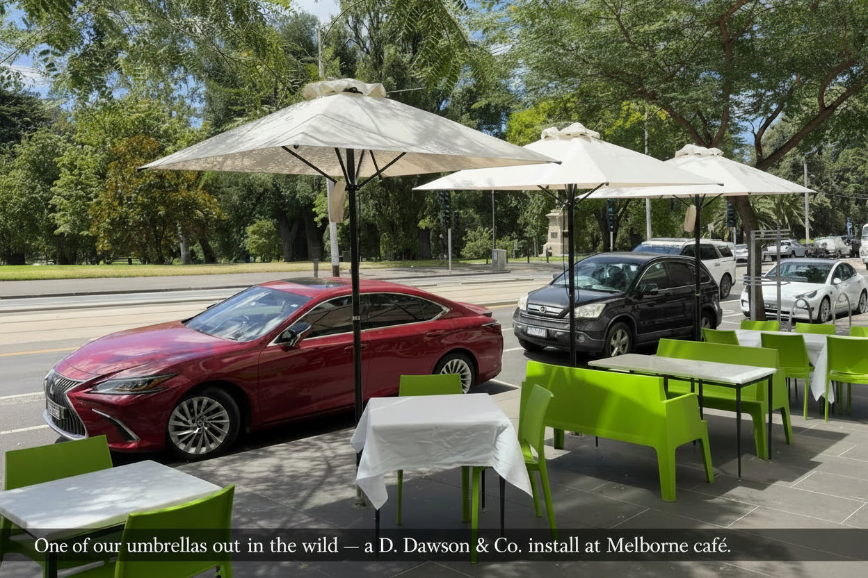 Outdoor seating area with tables, chairs, and umbrellas on a sidewalk with cars and trees in the background.